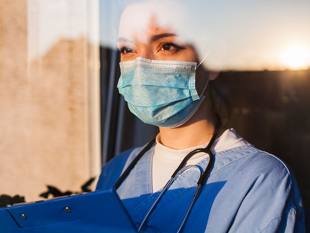 A woman wearing scrubs and a medical mask, with a stethoscope around her neck, and holding a clipboard, looks through a window.