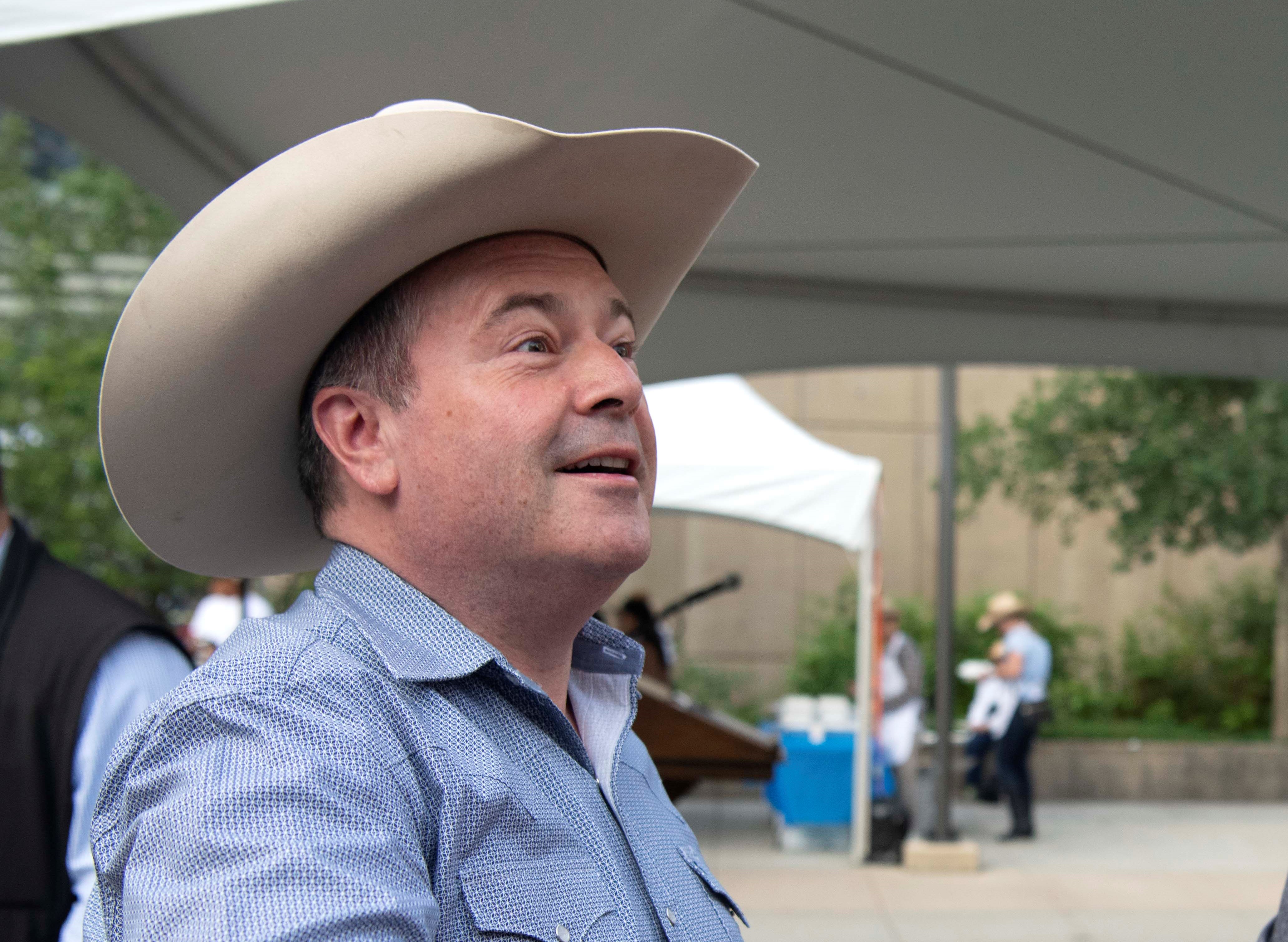 Seen from the side, a round-faced middle-aged man (Jason Kenney) wearing a white cowboy hat, smiles.