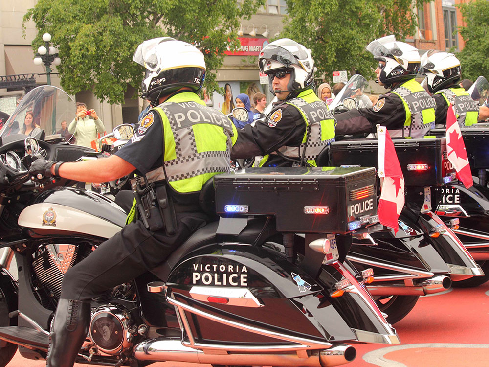 Police officers wear yellow safety vests as they ride motorcycles through downtown Victoria. The sides of their motorcycles are visible to the camera.