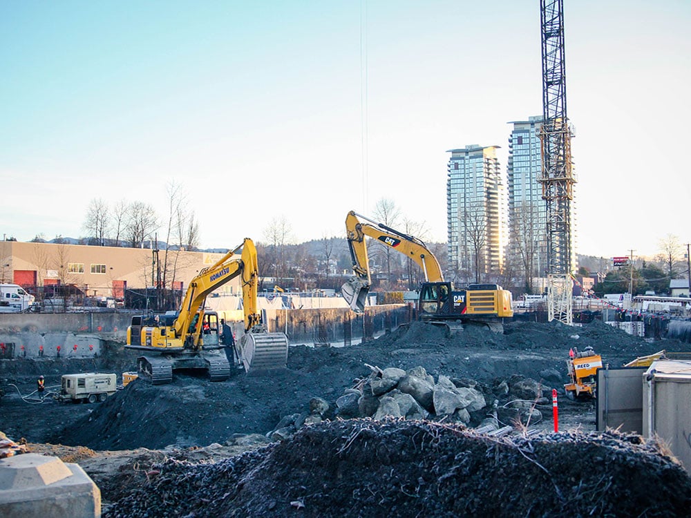 Two yellow excavators are working in a large construction site, with the base of a crane visible behind them. In the background are two residential towers.