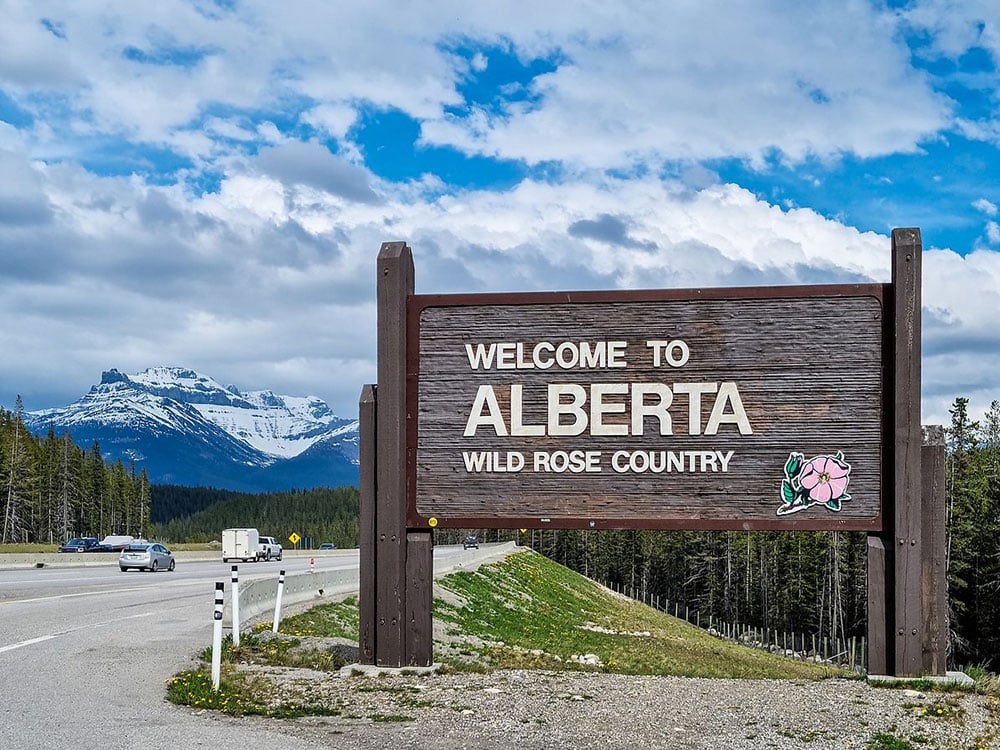 A highway sign says ‘Welcome to Alberta — Wild Rose Country.’ Coniferous trees line the four-lane highway. A snow-covered mountain rises against a background of blue skies and light clouds.