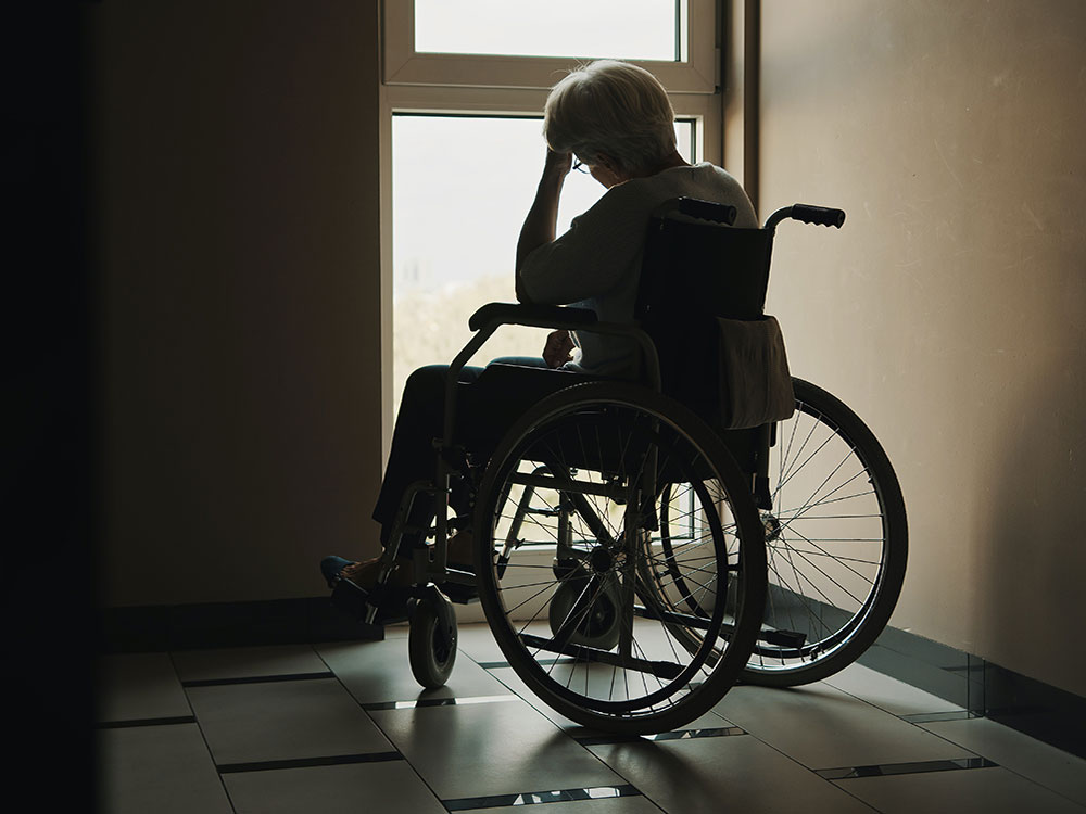 An older woman sits in a wheelchair in an empty hall. Her hand is raised to her head and her shoulders are slumped.