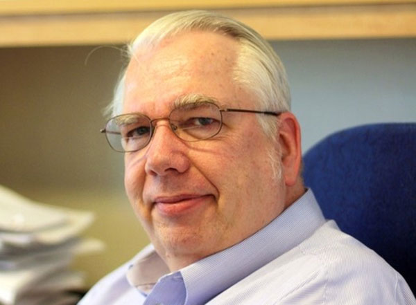 A man with silver hair and glasses, wearing a pale blue shirt with a button-down collar, looks at the camera.