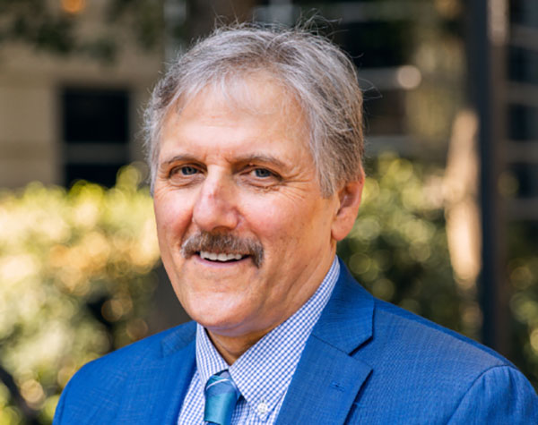 A man with greying hair and a moustache, wearing a blue suit, blue checked short and tie, smiles at the camera.