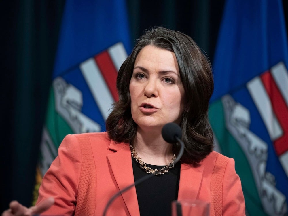 A dark haired woman wearing a peach jacket, black shirt and gold necklace stands in front of a microphone, with two Alberta flags behind.