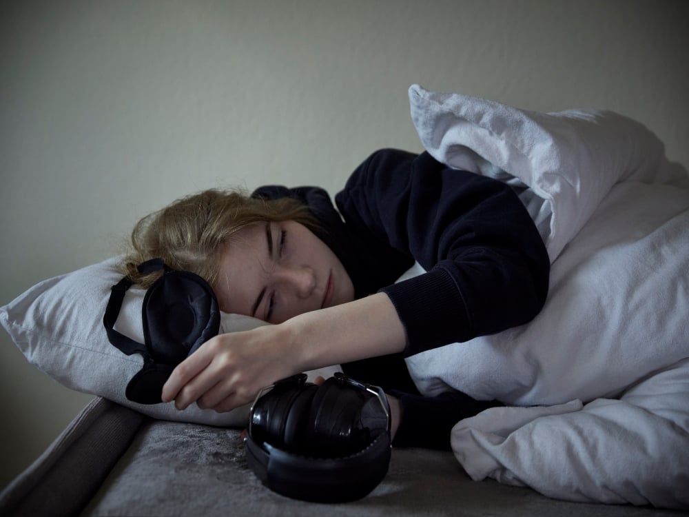 A young woman in a black hoodie lies in bed under a white duvet on a white pillow. She is holding a black eye mask. A pair of large black headphones is at her side. 