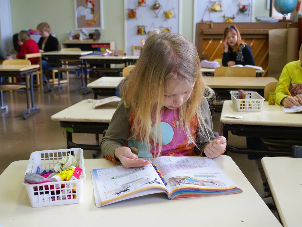 A young girl with blonde hair sits at a short white desk, looking at an open book. Her right palm is open as she is counting with her fingers. To her right is a small plastic basket of school supplies on her desk. Behind her are other empty desks. In the far background, students are seated and engaging in a lesson. 