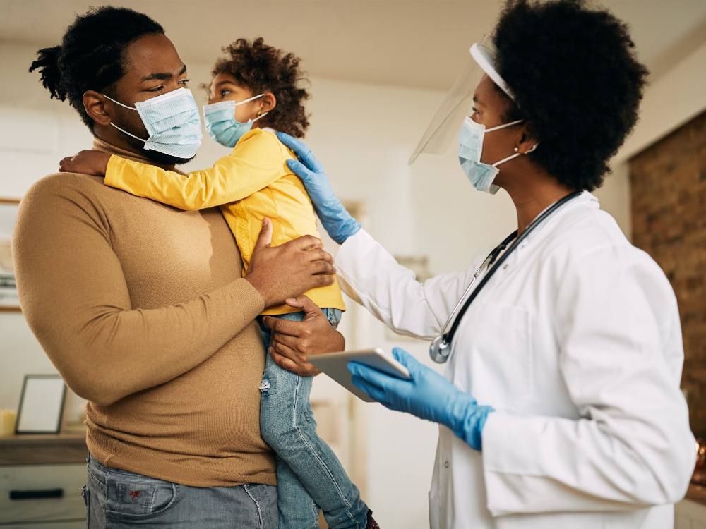 A physician in a white lab coat with a stethoscope around her neck stands to the right of the frame, placing her hand gently on the back of a young child in a yellow shirt to the left of the frame. The child is in their father’s arms; he is wearing a beige sweater and jeans. All three are in blue medical masks; the physician has a face shield.