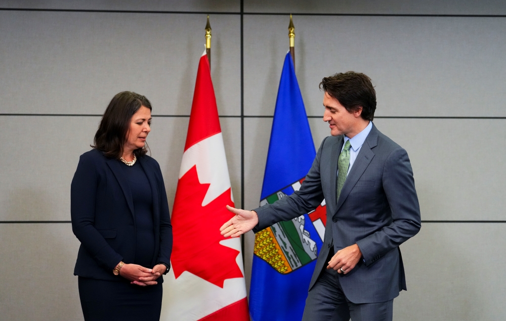 Danielle Smith, in a blue suit, stands with her hands folded at her waist as Justin Trudeau reaches out to shake her hand. He’s smiling. Smith is looking at his hand like it’s a dead fish. They stand in front of Canadian and Alberta flags.