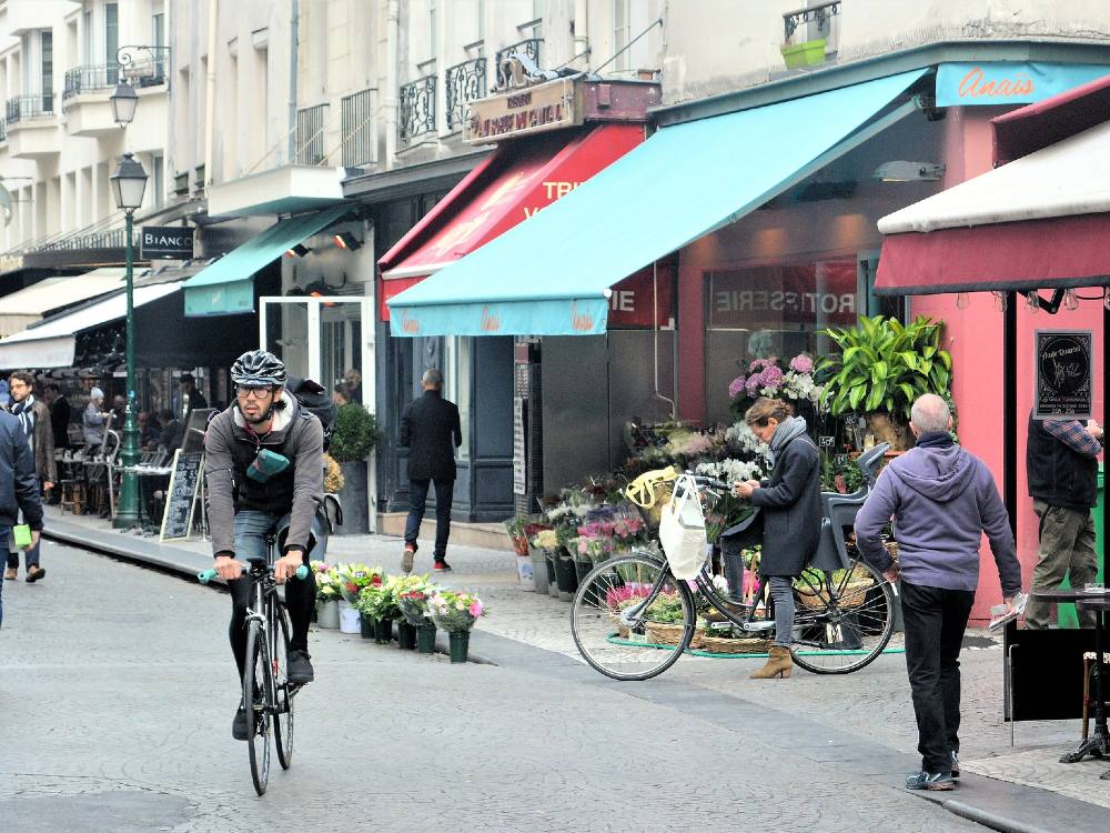 A city street in Paris. People are walking and cycling. There are storefronts at street level, and residences and offices above.
