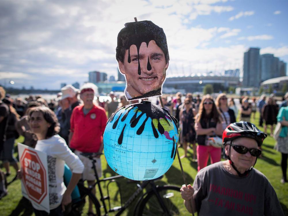 A protest placard bears the resemblance of Trudeau on top of a globe, with oil dripping down his head. The scene depicts a protest with many people walking together. 