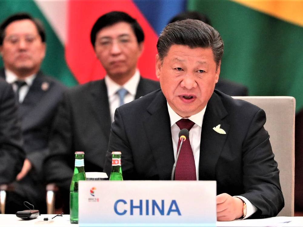 A middle-aged Chinese man, China’s leader Xi Jinping, wears a suit and sits at a table with a placard saying China, as two other standing Chinese men in suits look on from behind.
