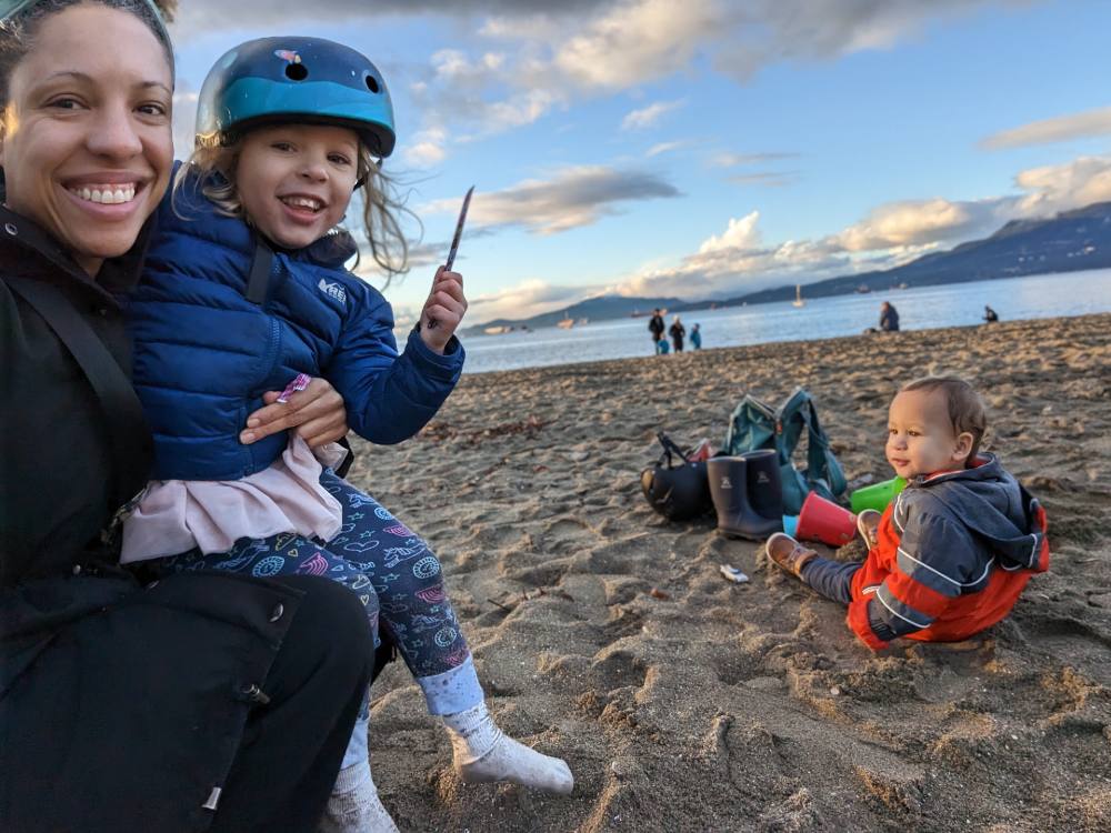 Brittany Hopkins is to the left of the frame, holding her young daughter. Hopkins is wearing a black jacket and her daughter is in a blue jacket with a blue bicycle helmet. Her toddler son sits on the sand in a grey and blue rain jacket. In the background is the ocean and blue sky.