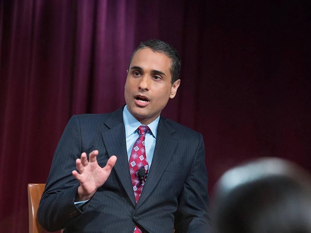 A person of colour in a suit speaks into a clipped-on mic on stage.