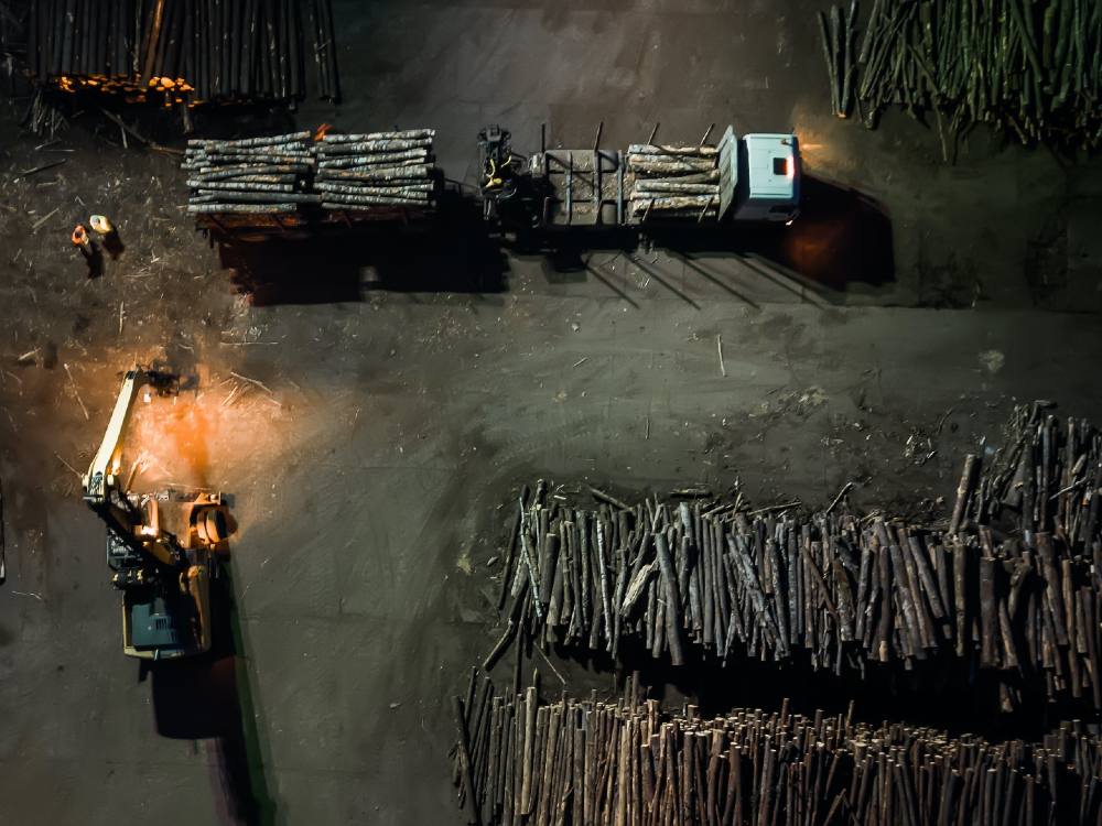 An overhead photo shows a truck at night in a large lot full of logs, being loaded under a bright light.