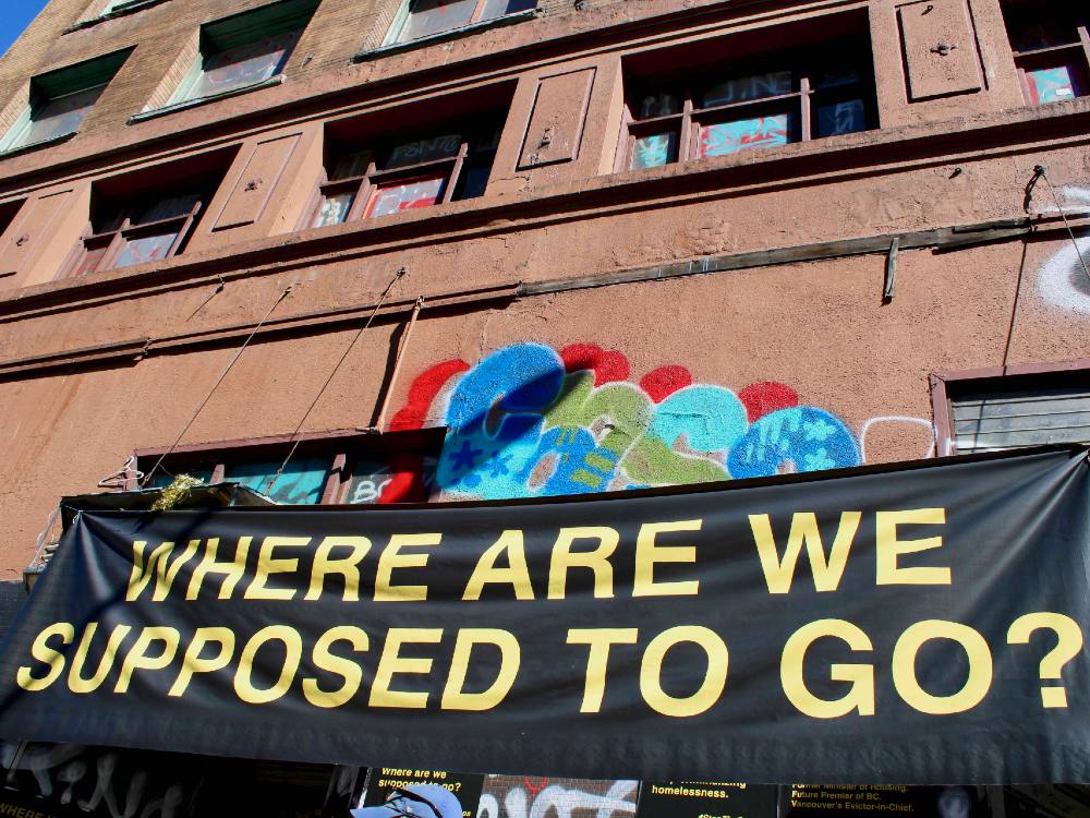 A horizontal black banner with yellow san-serif text reads “Where are we supposed to go?” in capital letters. The banner hangs across two doorways of a tall brown multi-storey building in the Downtown Eastside. Colourful graffiti can be seen behind the sign. It is a sunny day.