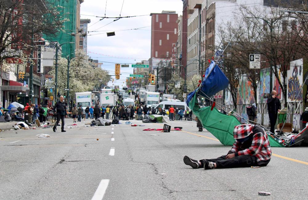 The westward view towards Columbia Street on East Hastings Street, Vancouver, on the day the police and city workers took apart a decampment on East Hastings. The street is empty with a person sitting slumped over in the middle of the road in black jeans and a red plaid jacket. In the background are lines of large white trucks behind a line of people.  