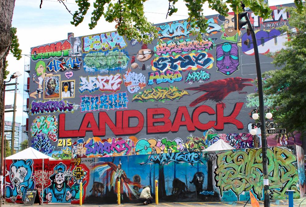 A colourful mural on a dark grey painted wall features the phrase “Land Back” in red capital letters. It is framed by numerous pieces of colourful artwork by different graffiti artists. 