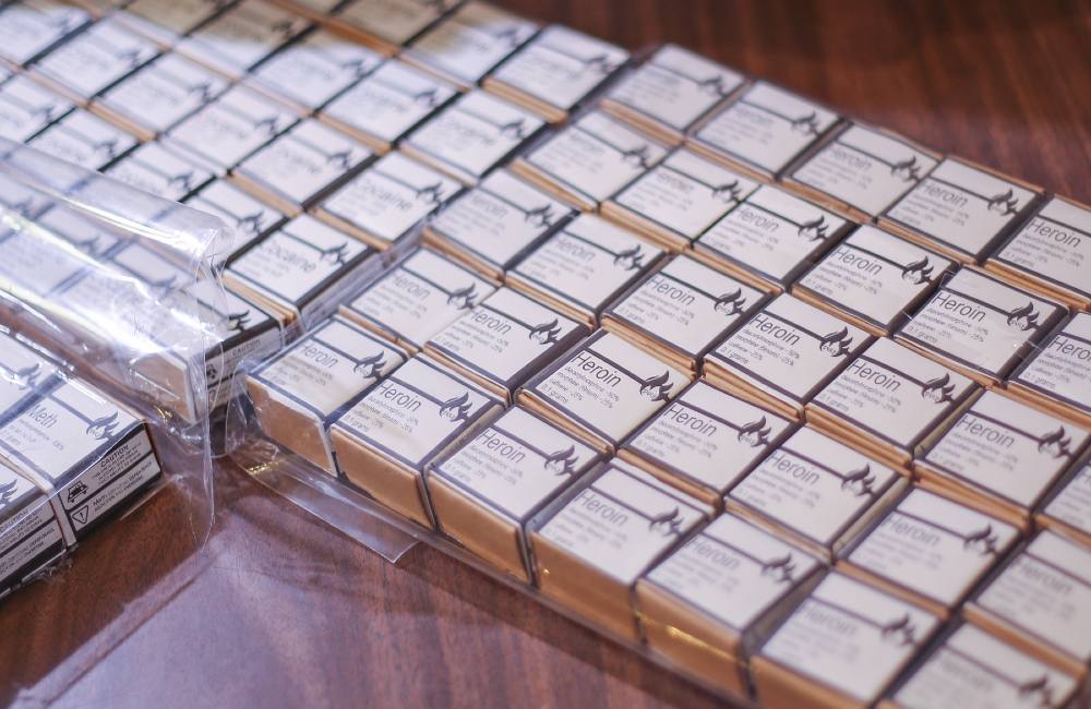 Numerous matchbox-sized boxes with white labels that read “Heroin” in san serif text line a wooden table. They are arranged in rows in large clear plastic bags with zip closures.