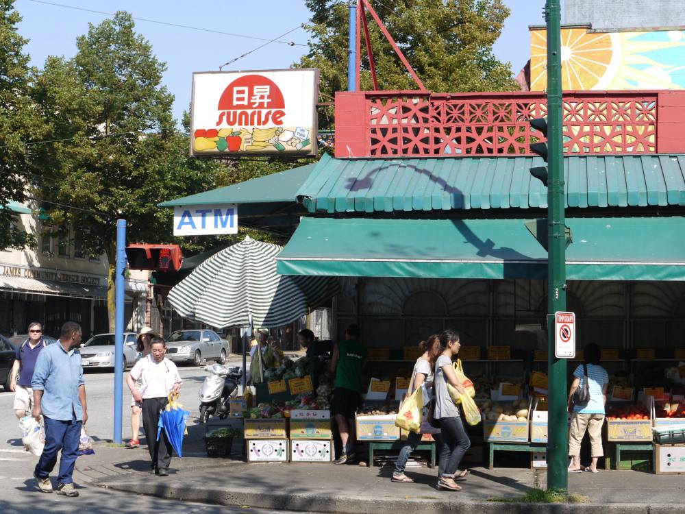 People shop at Sunrise Market on Powell Street on a sunny day. They are walking under a green awning where there are produce stalls. A square sign to the left of the frame reads ‘Sunrise’ in red text. 