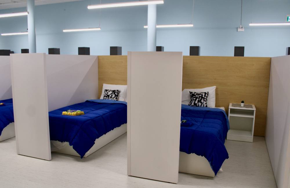 Two single beds with royal blue bedspreads are separated by low wooden partitions. Long white neon lights hang from the ceilings against light blue painted walls.