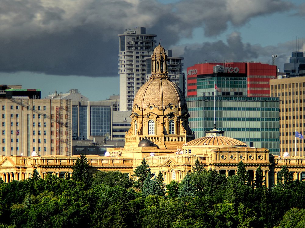 The Alberta Legislature building in Edmonton glows in the sunshine. Trees are in the foreground, while numerous downtown buildings are seen off in the distance.