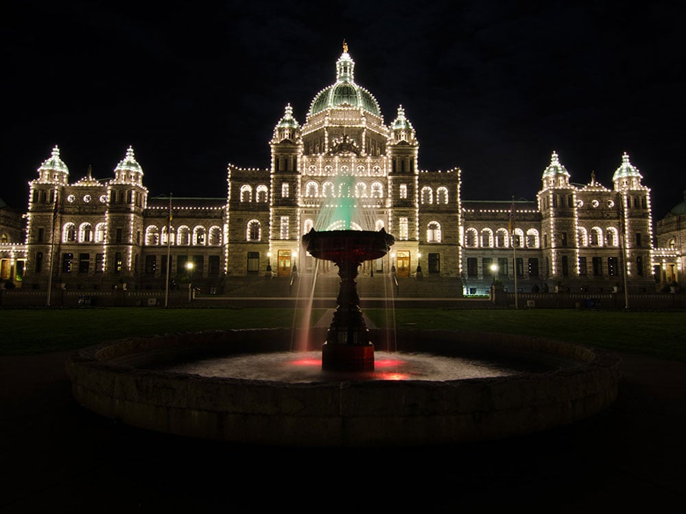 A night shot of the BC Legislature building outlined with lights and a lit fountain in the foreground.