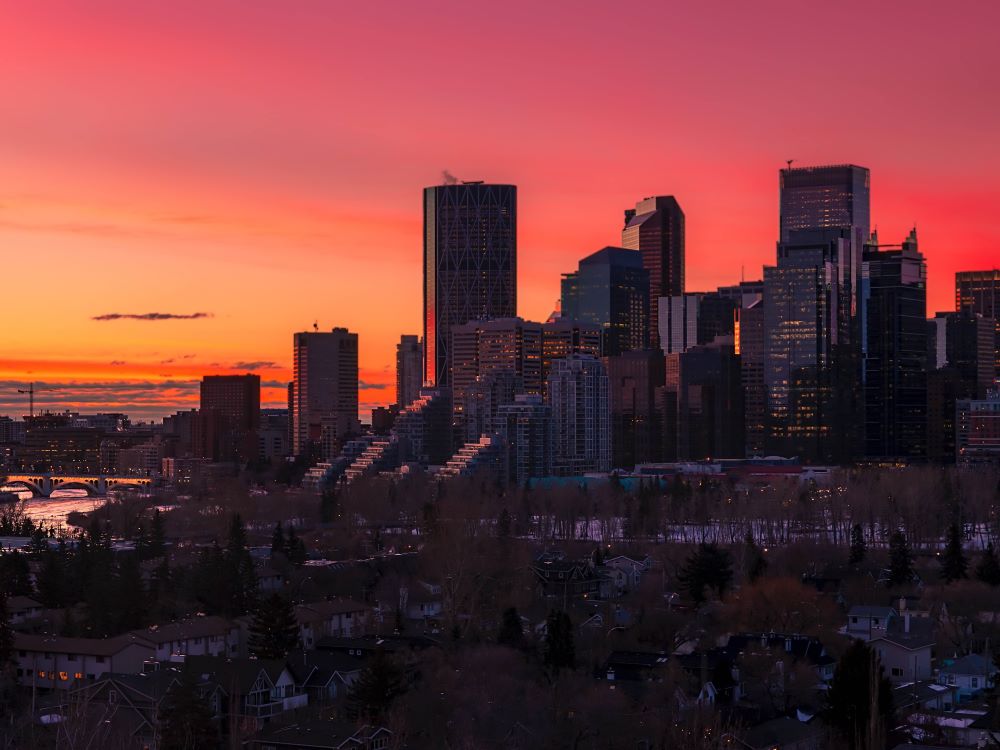 The downtown Calgary skyline at sunrise features an array of high-rise office buildings against a pink and orange sky.