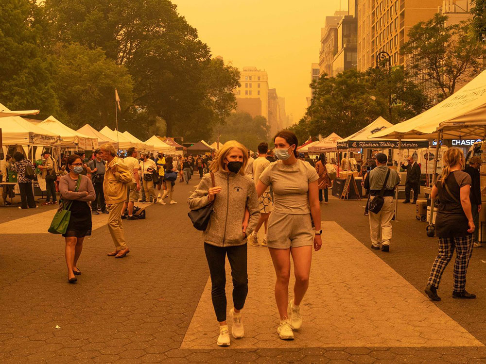 Two women in grey and black athletic apparel walk down the middle of the frame on a tiled concrete walkway. They are both wearing medical masks, as are other patrons amidst the farmers’ market stalls on both sides of them. The photo has a hazy orange hue from wildfire smoke.