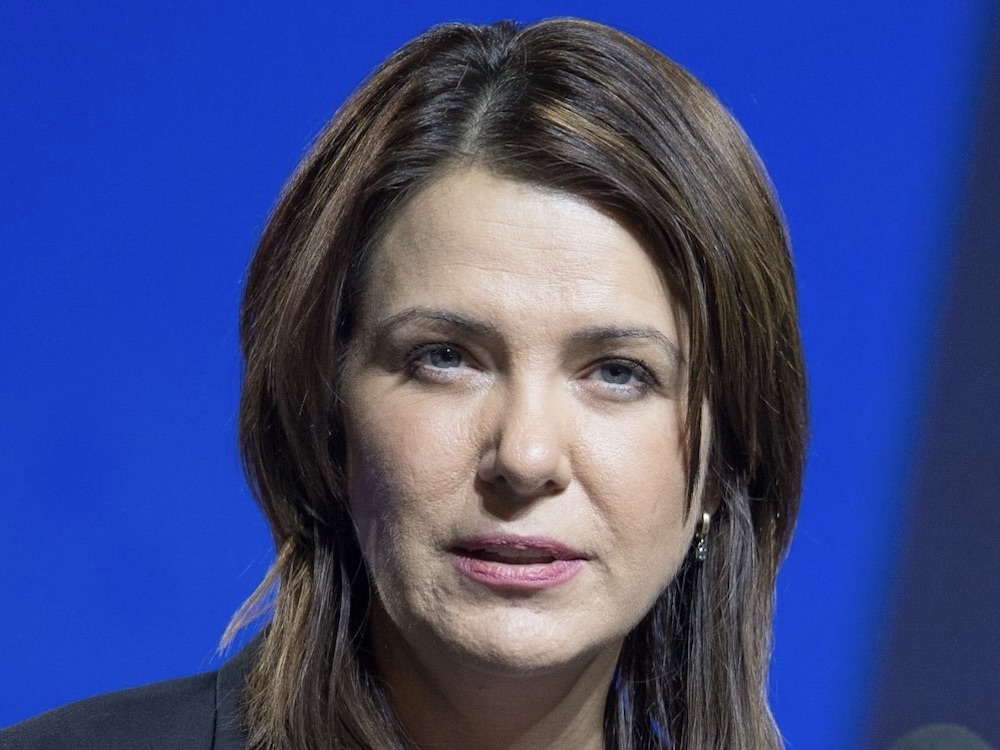 A close-up photo of Alberta Premier Danielle Smith’s face on a royal blue background.