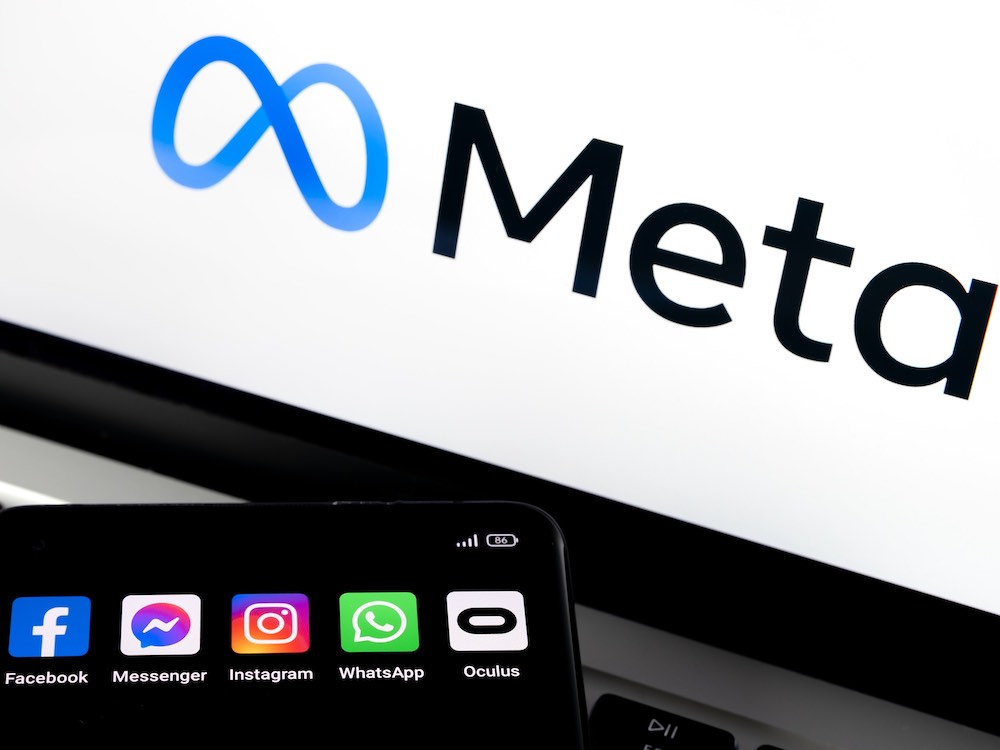 A black smartphone is held up at the bottom of the frame against a laptop screen that displays the logo for Meta against a white background. The smartphone displays apps for Facebook, Facebook messenger and Instagram, which are owned by Meta. Other apps on the phone include WhatsApp and Oculus.