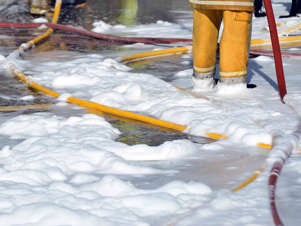 A firefighter in a yellow uniform, shot from the waist down, is up to their ankles in white, fluffy firefighting foam. The foam covers a street lined with yellow firefighter hoses. 