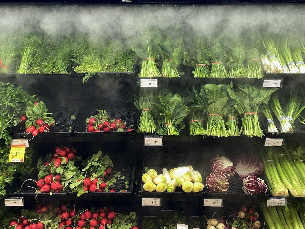 Grocery shelves are packed with produce, while a mister emits a fine spray.