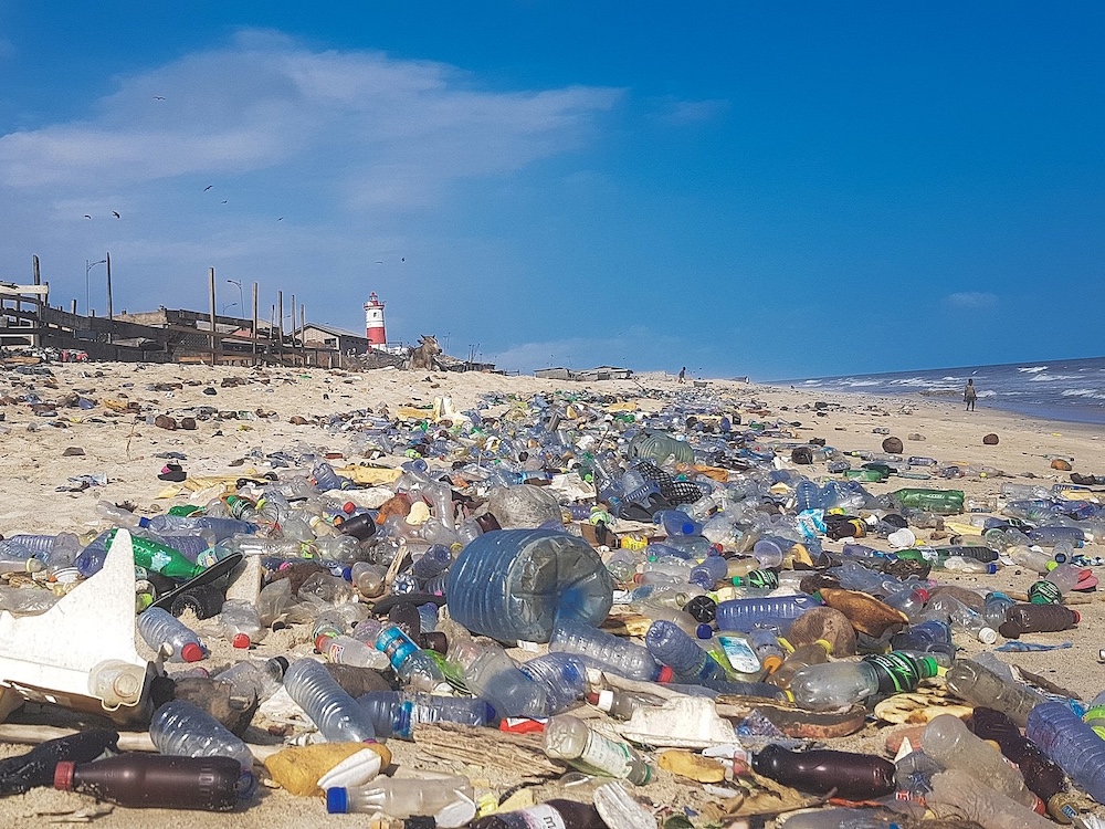 Plastic waste is strewn across a beach under a blue sky, with a lighthouse in the background.