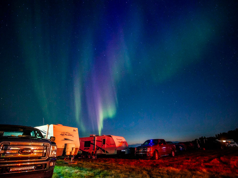 Trucks and camping trailers are parked in a field under stars and the northern lights.