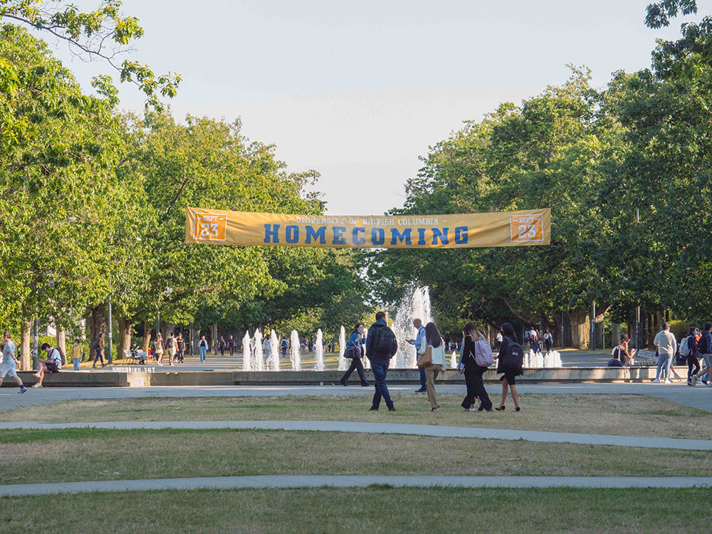 Young adults walk across a tree-lined plaza with a water fountain in the middle. Their backs are turned to the camera and they are wearing backpacks. A yellow banner with the word “Homecoming” in blue varsity typeface hangs over the fountain between the trees.
