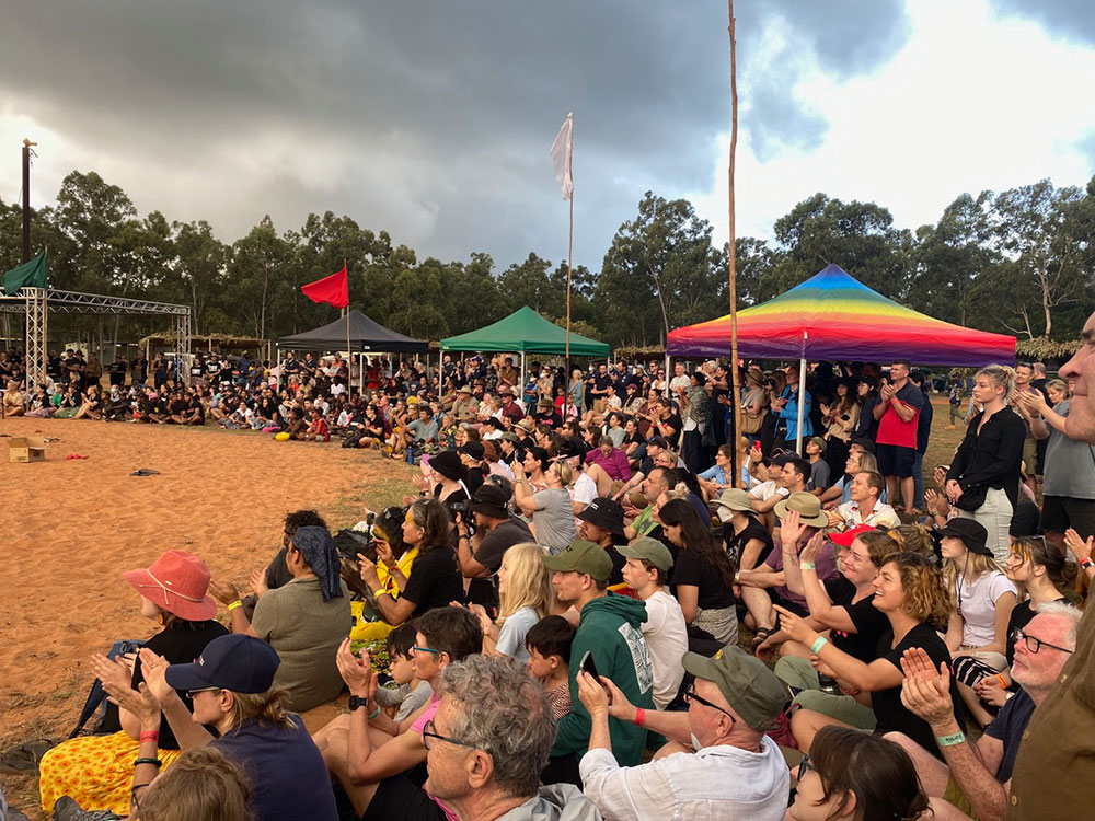 A group of people sit on the sand in a semi-circle surrounded by tents and a stage, watching a performance at the Garma Festival, Australia’s largest Indigenous festival.