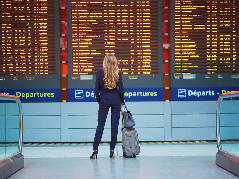 A young woman with long blonde hair stands in front of a digital airport schedule with writing in yellow on a black background. She is wearing dark blue and a carry-on bag and a grey rolling suitcase rest on her right.