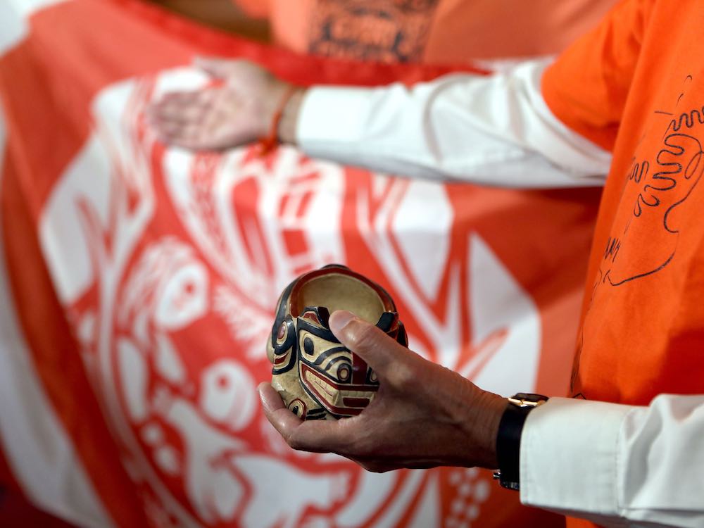 In sharp focus in the foreground, a hand with white sleeves under an orange T-shirt holds a ceremonial wooden water bowl decorated with black and red Indigenous carvings. In soft focus in the background, the orange and white Survivors’ Flag in honour of residential school survivors and those who died in the schools.