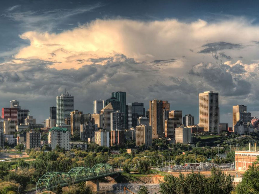 A sky with spectacular clouds over the skyline of Edmonton seen from a distance, with low-rise buildings and foliage in the foreground.