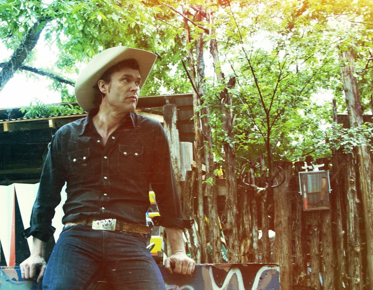 A man in a cowboy hat, black button-up shirt, jeans and a large gold belt buckle looks off to his left. Behind him can be seen a lean-to, rough-hewn fence and some greenery.