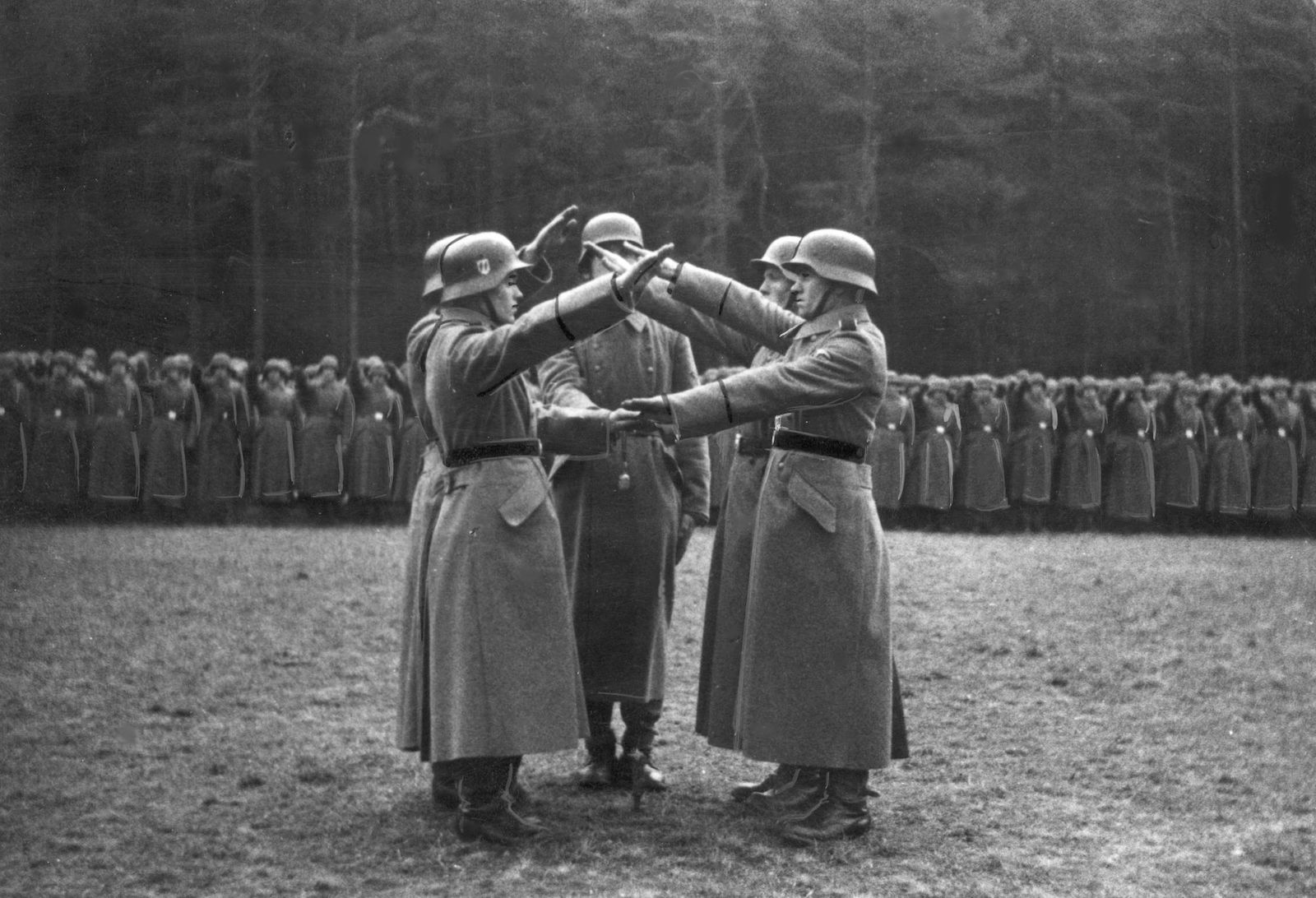 An old black and white photo shows a group of five soldiers wearing long coats and bucket helmets giving the Nazi salute in the middle of a grass field. In the background a legion of similarly dressed soldiers can be seen also giving the salute.