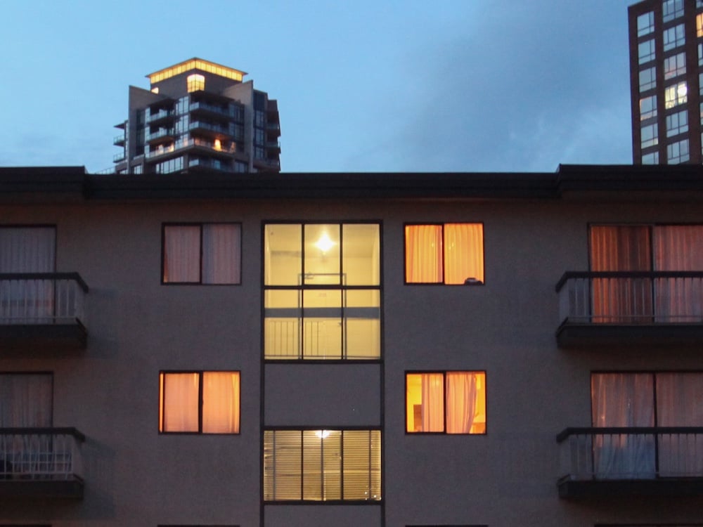 In the foreground, a wide two-storey apartment building stands at dusk. The building is dark and studded with warm interior light from some of the windows. Behind the two-storey building, taller, newer highrise residential buildings stand against a darkening cloudy sky.