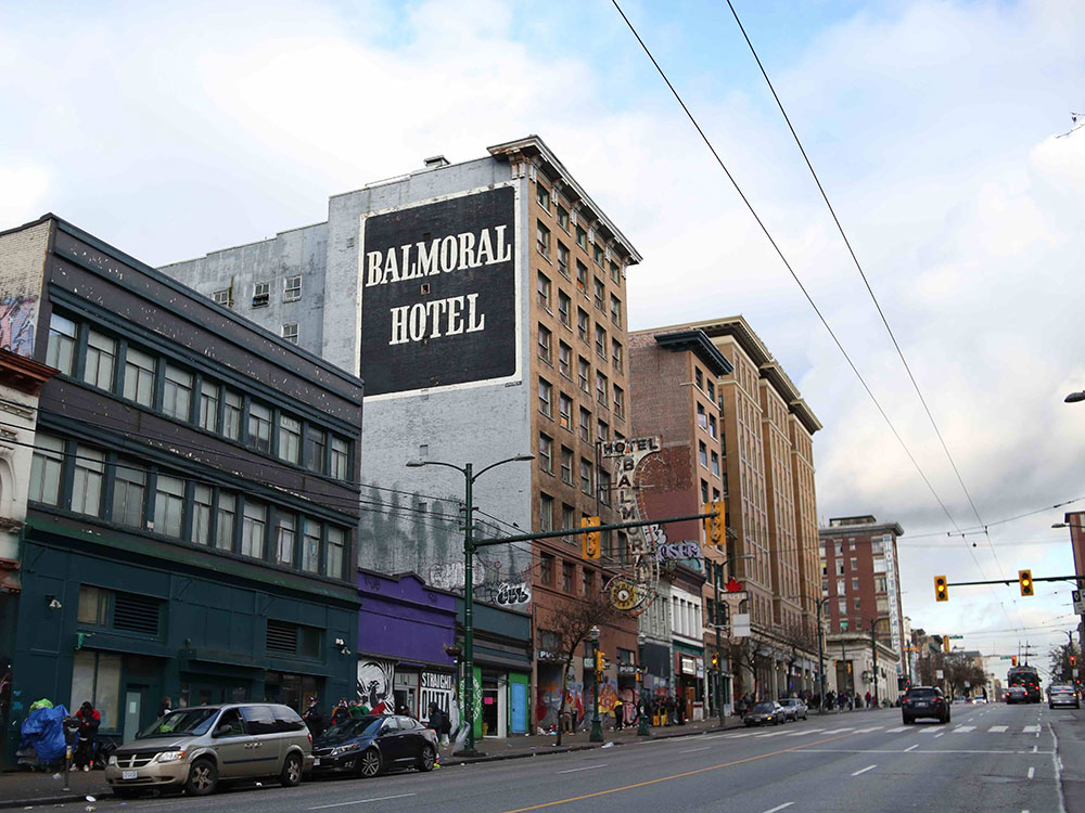 The view of the northeast corner of East Hastings Street at Main in Vancouver. Fluffy white clouds obscure a cold blue sky. A tall brown brick building is painted grey on one side with the words ‘Balmoral Hotel’ in white all-caps serif script against a black squarish background. It is surrounded by other brick buildings of varying height. A few vehicles are visible parked and on the road. The sidewalk reveals some signs of street activity, but the people are out of focus.