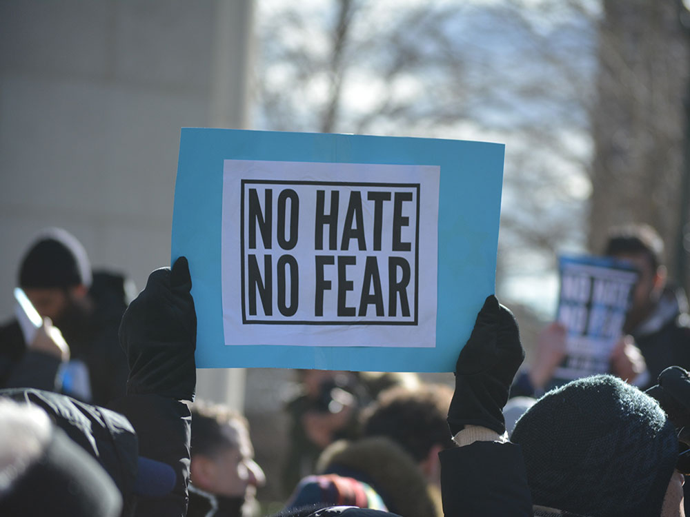 Protesters are wearing cold-weather clothes. In the centre of a frame, a protester holds up a sign saying, ‘No Hate, No Fear.’