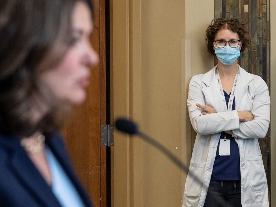 Brown-haired Premier Danielle Smith’s face is seen from the side and out of focus. Standing against a wall looking on, and in focus, is a woman with glasses and curly hair, wearing a medical mask, with arms folded and her middle finger extended in a non-demonstrative way.