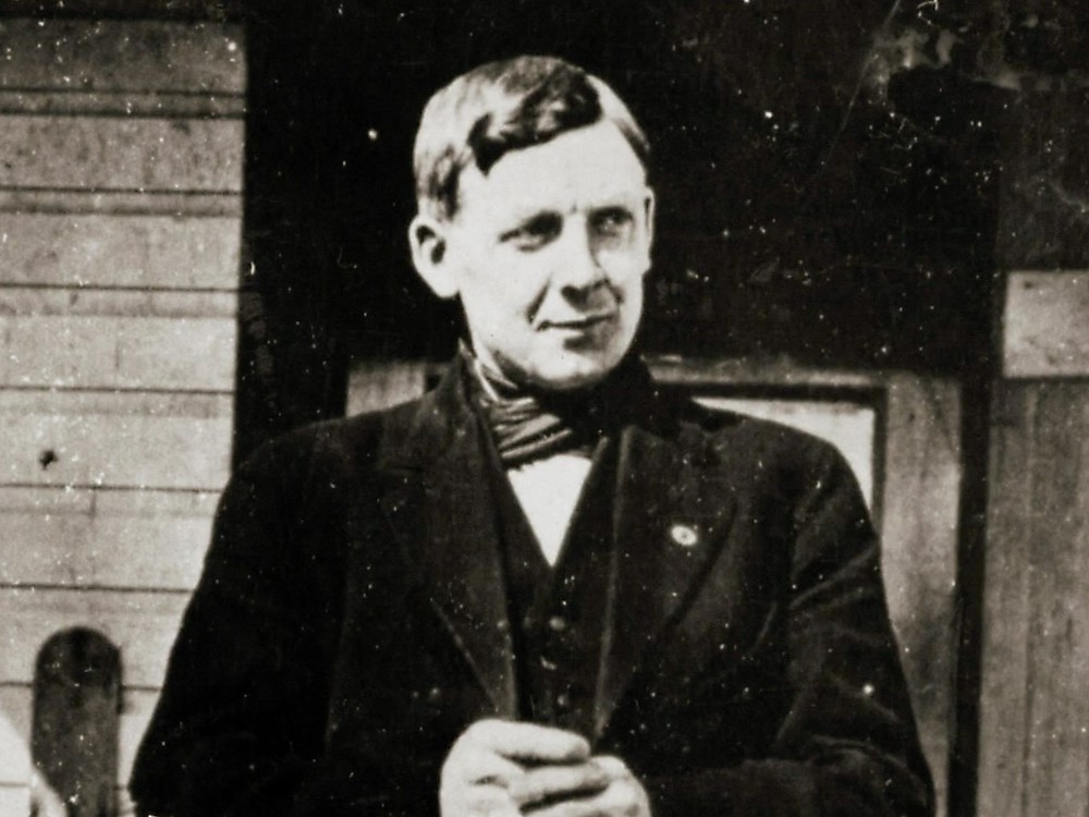 A black and white photo of a white man in his 30s wearing a black coat and vest, round button on his lapel and a scarf around his neck.