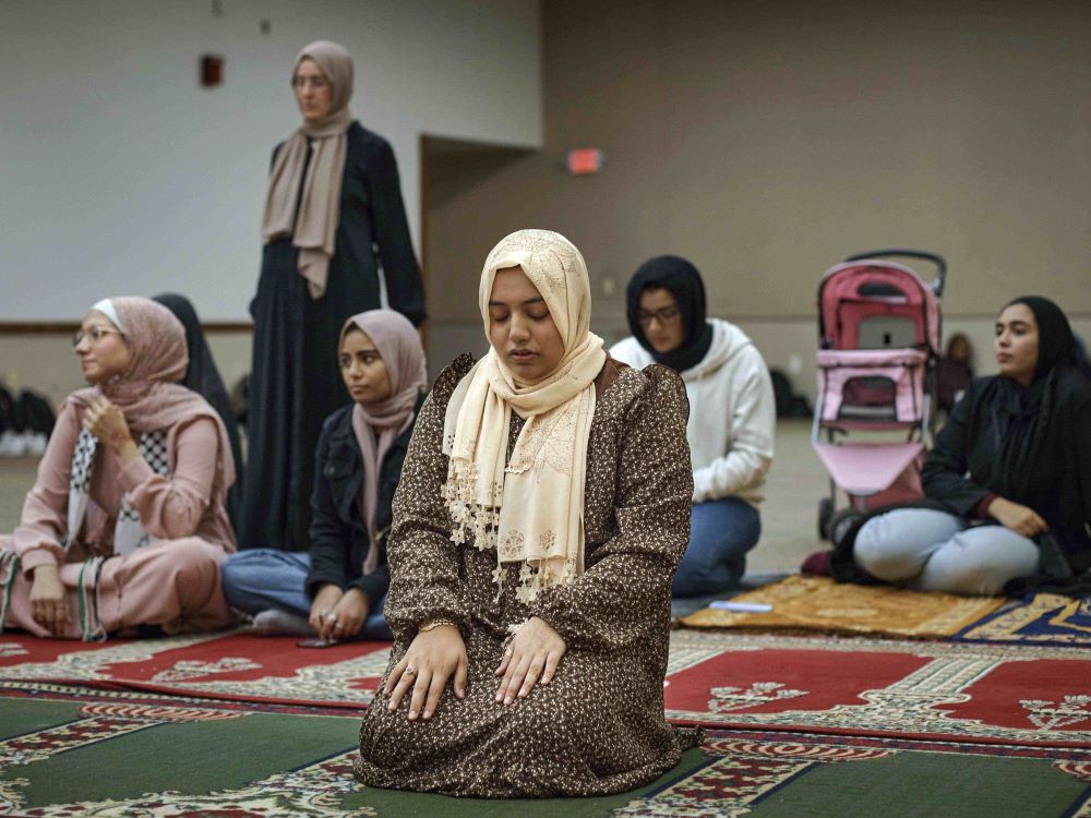 A group of young women sit on a collection of thick colourful rugs in a room with beige walls. They are wearing hijabs, head coverings worn by some Muslim women. The woman in the foreground is wearing a light beige head covering and a brown floral long-sleeved dress. She kneels with both hands on her thighs, looking down.