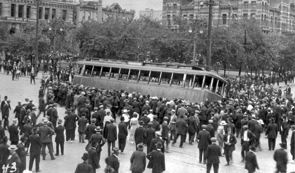 A black and white photo shows a large crowd tipping a streetcar.