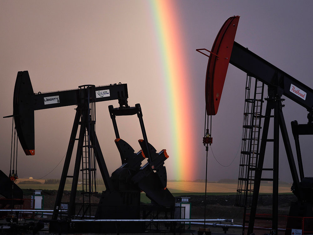 Two large oil well pumps stand on the prairie, with a brilliant rainbow descending to the ground behind them in front of grey skies.